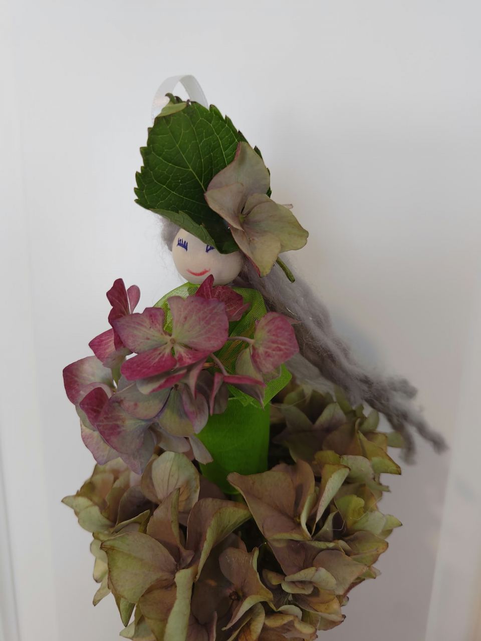 Selbstgebastelte Herbstfigur: Holzkopf mit grünem Blatt als Hut, grauen Wollhaaren, grünem Kleid und einem Rock aus verblühten Blütendolden