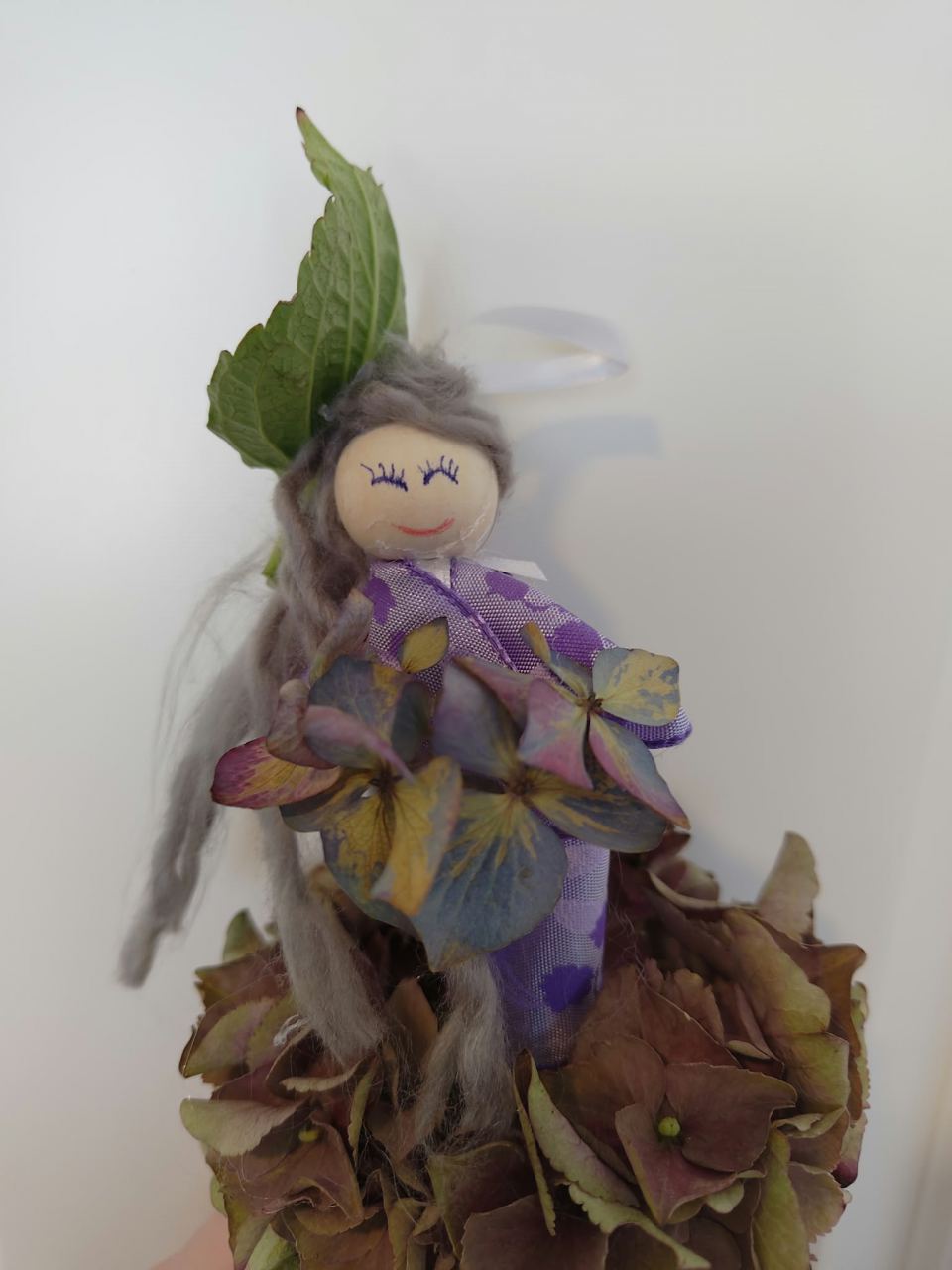 Selbstgebastelte Herbstfigur in lila Kleid mit weißer Schleife, grauen Wollhaaren und einem Rock aus braunen Blütendolden