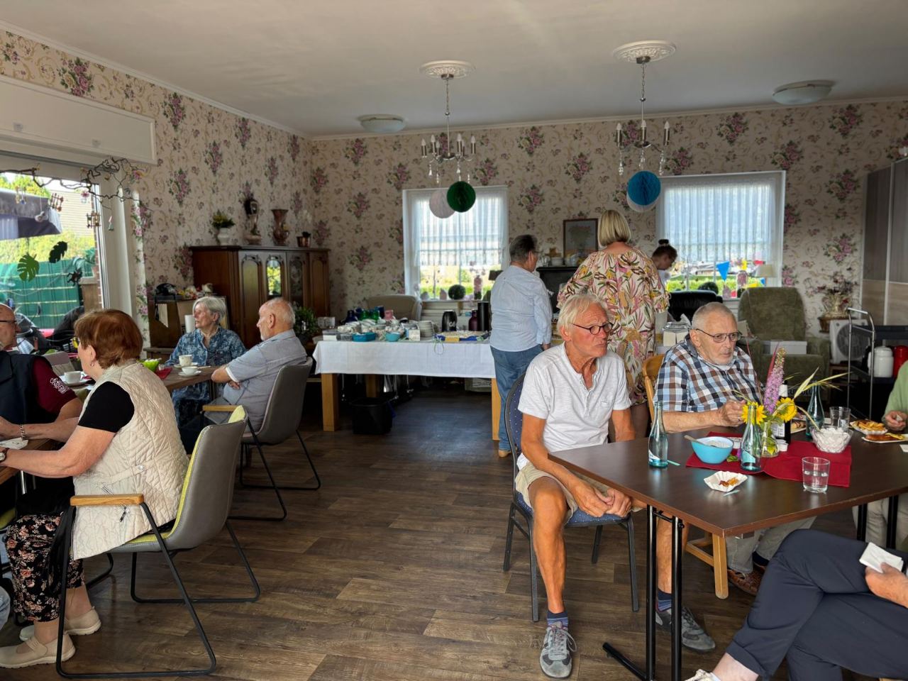 Gäste sitzen beim Sommerfest drinnen am Kaffeetisch, im Hintergrund der Blick in den Garten